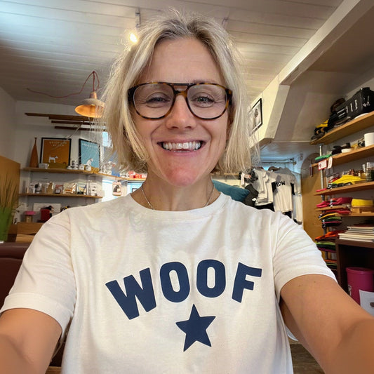 A smiling woman with short blonde hair and tortoiseshell glasses takes a selfie while wearing a white t-shirt with the word 'WOOF' and a star printed in navy blue. The background shows a cozy, colorful boutique interior.