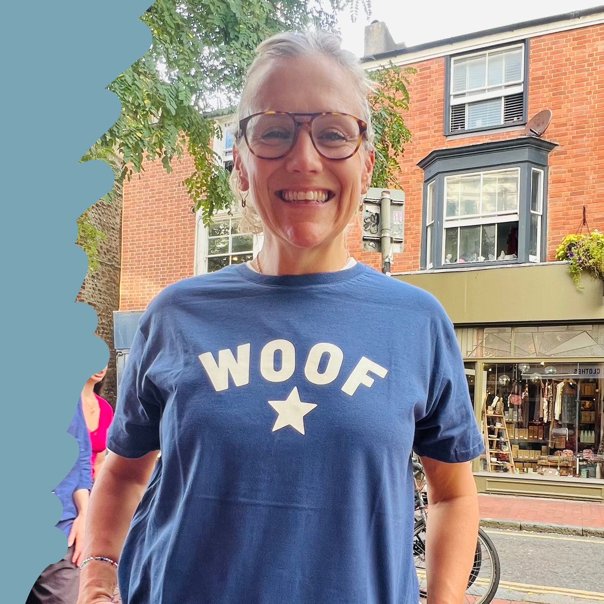 A woman wearing a navy blue t-shirt with 'WOOF' and a star graphic printed in white, paired with dark wide-leg jeans, standing on a brick sidewalk in an outdoor street setting.